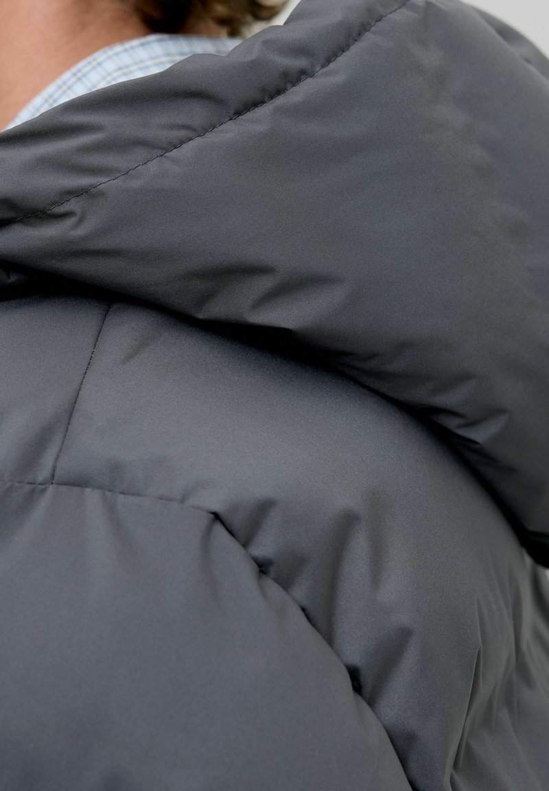 Primo piano di una persona che indossa una giacca imbottita grigio scuro con un ampio colletto, che mostra parzialmente una camicia a quadri blu sottostante.