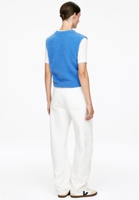 Blauer ärmelloser Pullover mit einer weichen Textur, über einem weißen kurzärmeligen Hemd, kombiniert mit weißen weit geschnittenen Hosen und Turnschuhen.