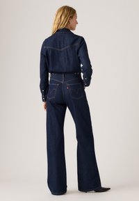 Camisa de ganga azul escura e jeans de perna larga com costura tonal. Apresenta pala nas costas e dois bolsos traseiros, com sapatos pretos de bico visíveis.