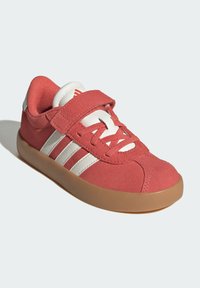 červené semišové tenisky s bielymi pruhmi, gumová podrážka a suchý zips cez šnúrky. Zaoblená špica a polstrovaný golier.