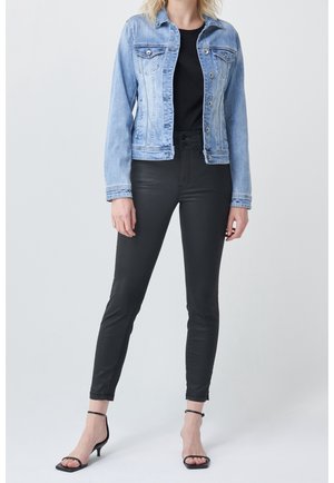 Femme portant une veste en jean bleu clair, un haut noir, un pantalon noir ajusté et des sandales à talons noires avec bride à la cheville, debout devant un fond uni.