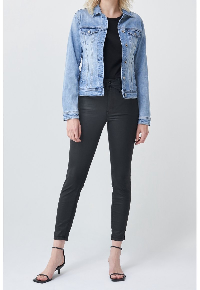 Femme portant une veste en jean bleu clair, un haut noir, un pantalon noir ajusté et des sandales à talons noires avec bride à la cheville, debout devant un fond uni.