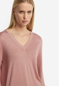 Jasnoróżowy sweter z dekoltem w serek i długimi rękawami, o gładkiej teksturze i subtelnym połysku, wykonany z miękkiego materiału. Bez wyraźnych wzorów ani akcentów.