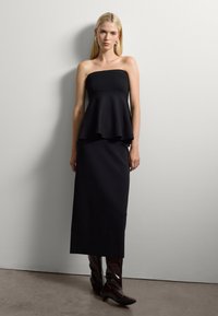 Frau mit langen blonden Haaren trägt ein strapless schwarzes Peplum-Oberteil, einen schwarzen Midi-Rock, kastanienbraune Stiefeletten mit Absatz und silberne Creolen vor einer grauen Wand.