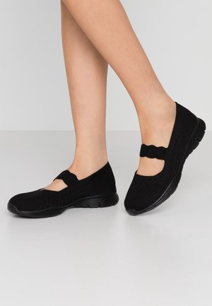Damskie nogi w czarnych dzianinowych, codziennych butach w stylu Mary Jane z elastycznymi paskami, na jednolitym jasnym tle.