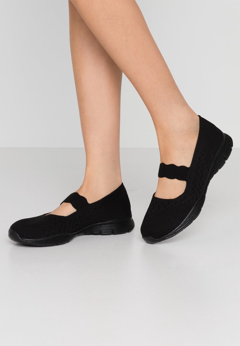 Jambes féminines portant des chaussures casual en maille noire style Mary Jane avec des sangles élastiques, sur un fond clair uni.