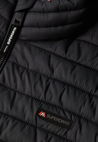 Chaqueta negra acolchada con costuras horizontales, etiqueta de la marca Superdry® visible y tirador de cremallera en la superficie de tela suave.