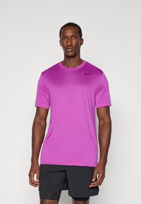 T-Shirt mit kurzen Ärmeln aus leuchtend lila Stoff, ausgestattet mit einem runden Ausschnitt und dem schwarzen Nike-Logo auf der Brust. Tragend mit schwarzen Shorts.