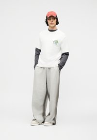 Νέος άνθρωπος που φοράει κόκκινο καπέλο, λευκό κοντομάνικο μπλουζάκι με μικρό γραφικό, μακριά γκρι μανίκια από κάτω, άνετο γκρι παντελόνι και αθλητικά παπούτσια.