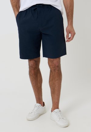 Mand iført marineblå shorts med snøre og hvide sneakers, stående med hænder afslappet ned langs siderne mod en ensfarvet baggrund.