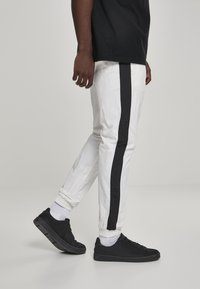 Hvite joggebukser med svarte side-striper, elastisk midje og mansjetter, kombinert med svarte sneakers og hvite sokker, sett fra siden.