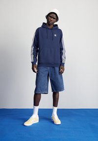 Marineblå hoodie med vit Adidas-logga och tre vita ränder på ärmarna, ihop med denimshorts och gula sneakers, står på en blå yta.
