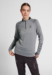Maglia sportiva grigia a maniche lunghe con colletto a mezza zip, tessuto testurizzato e accenti scuri; presenta un logo e un motivo a zigzag sulle maniche.