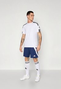 Weißes Sportshirt mit schwarzen Streifen, kombiniert mit marineblauen Shorts mit weißem Akzent und Logo. Weiße Socken und Fußballschuhe vervollständigen das Outfit.