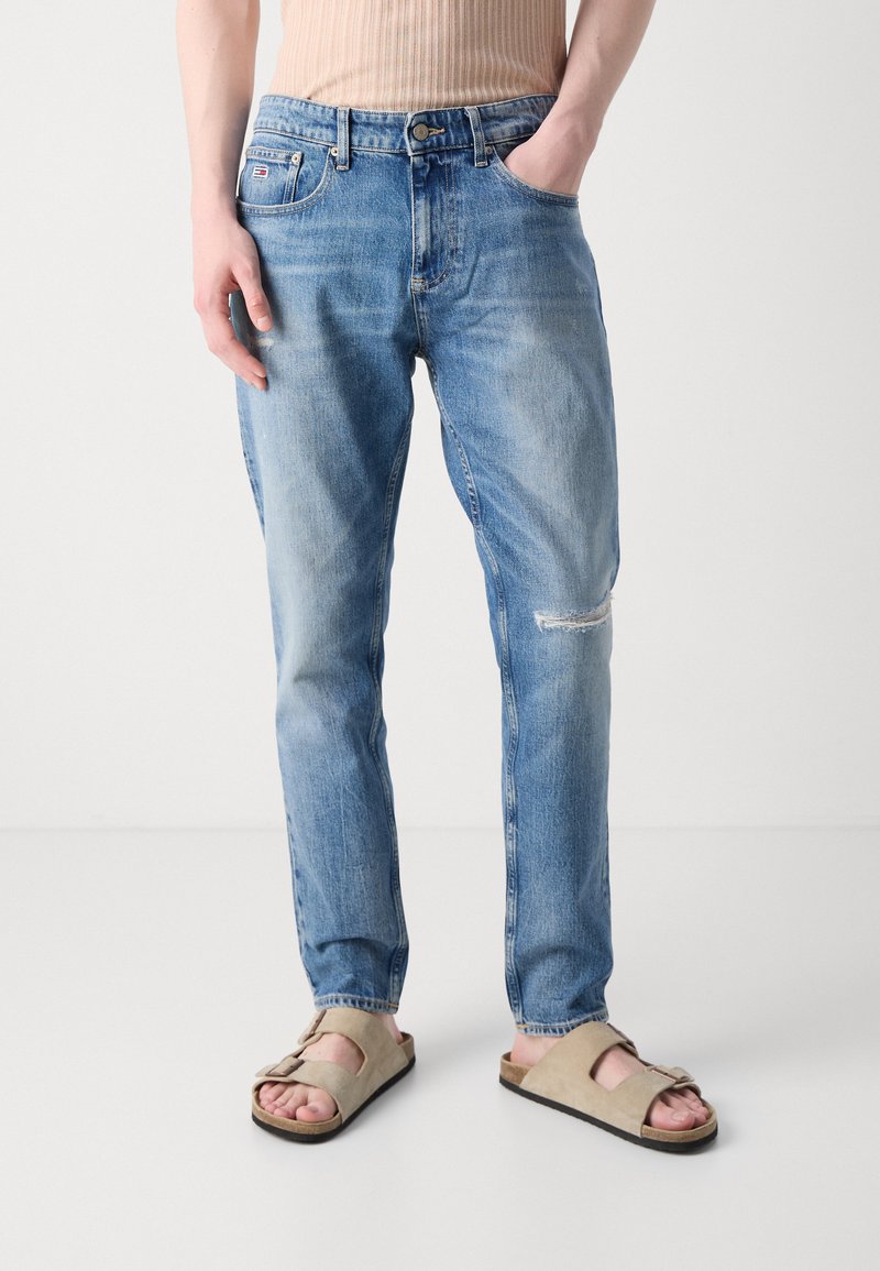 Helle blaue Jeans mit lockerer Passform, die ein kleines Rissdetail am Knie und ein klassisches Fünf-Taschen-Design aufweisen. Tragen mit beigen Sandalen.
