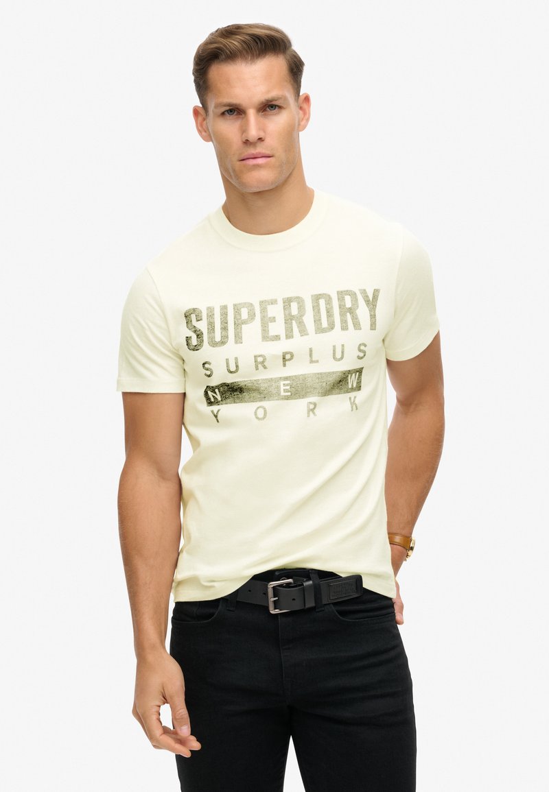 Lysegul bomuld t-shirt med fed, mørk grafisk tekst, der står "SUPERDRY SURPLUS NEW YORK." Korte ærmer og rund halsudskæring.