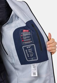 L'interno della giacca STX™ presenta una tasca blu navy con etichetta RECCO, dettagli impermeabili, traspiranti e antivento; tessuto grigio chiaro.