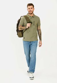 Khaki poloshirt met een ritszak, gedragen met lichtblauwe slim-fit jeans. Het model draagt een donkergroene rugzak met meerdere zakken.