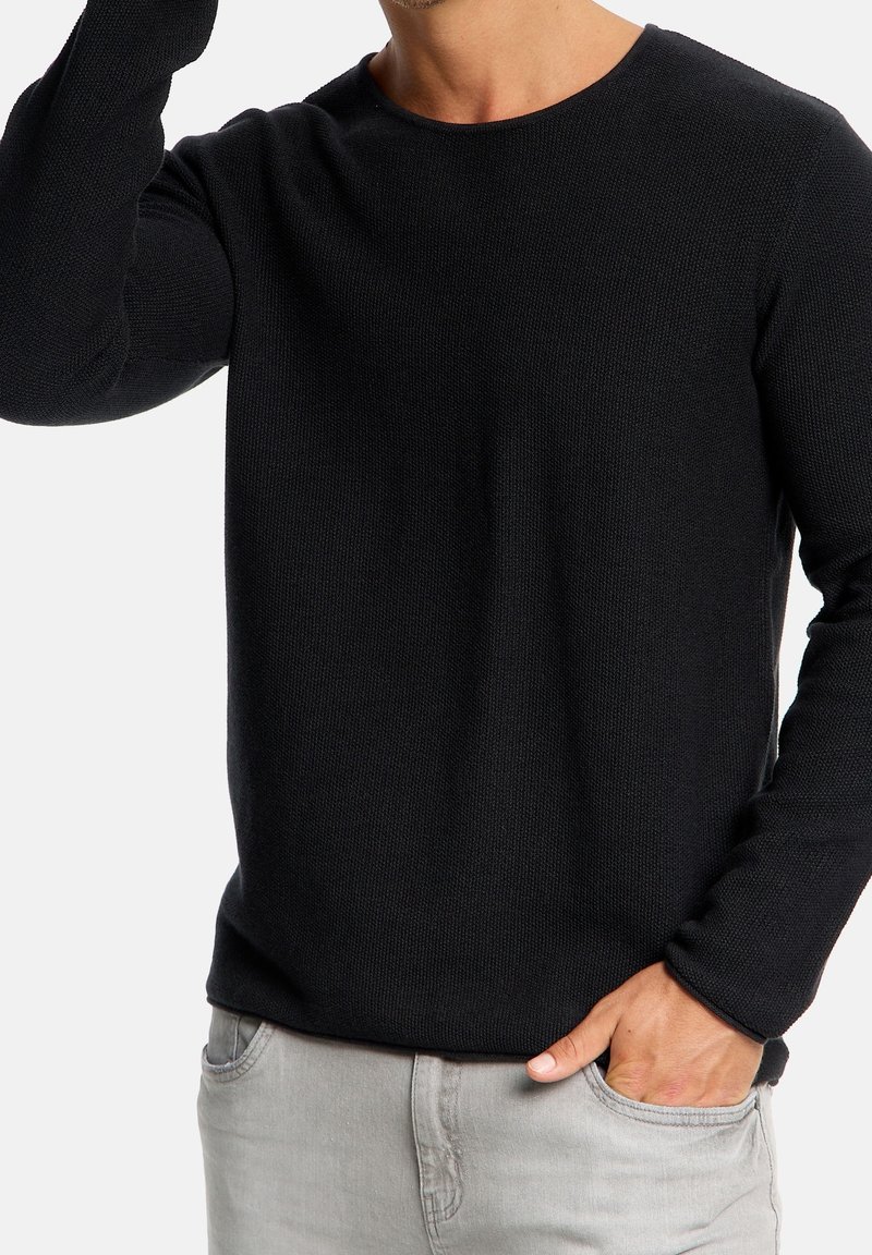 Pull en tricot noir à manches longues avec un col rond. Présente un tissu texturé et une silhouette ajustée. Associé à un pantalon gris clair.