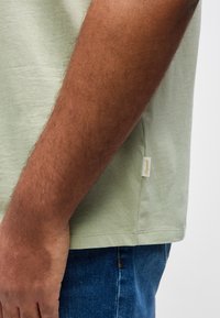 Πλάγια όψη ενός ατόμου που φοράει ανοιχτοπράσινο μπλουζάκι και μπλε τζιν, εμφανίζοντας το χέρι και μέρος του κορμού με μια μικρή ετικέτα ρούχο υ.