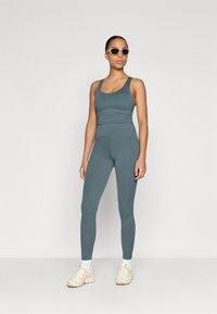 Modelis val wearing fitted, tirkīza krāsas treniņu topu un legingus, apvienojot tos ar bēšiem sporta apaviem, uzsverot gludu, elastīgu audumu un minimālu šuvju skaitu.