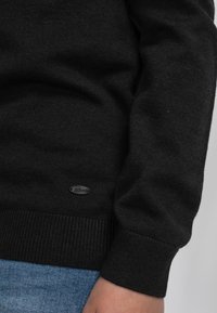 Schwarzer Strickpullover mit gerippten Bündchen und Saum, versehen mit einem kleinen grauen Logo-Patch an der Unterseite. Getragen über blauer Jeans.