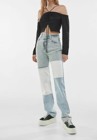 Top corto negro sin mangas con fruncidos, combinado con jeans de mezclilla de patchwork en azul claro que presentan secciones de tela blanca y zapatillas de plataforma.