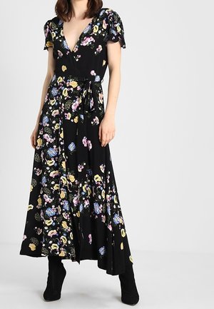 Kvinde iført en sort floral omslagskjole med korte ærmer og sorte ankelstøvler, stående mod en ensfarvet lys baggrund.