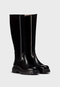 Las botas de cuero negras hasta la rodilla presentan un diseño elegante con punta redonda y una suela de goma texturizada. Detalle notable de cremallera lateral.