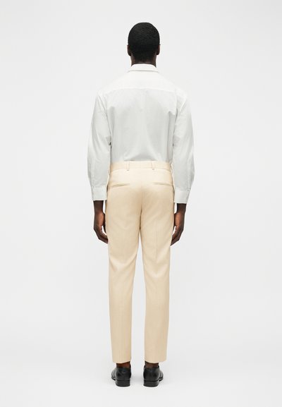 Άνδρας που φοράει λευκό πουκάμισο με μακριά μανίκια, μπεζ προσαρμοσμένο παντελόνι και μαύρα επίσημα παπούτσια, στέκεται με την πλάτη προς το μέρος μας μπροστά σε απλό λευκό φόντο.