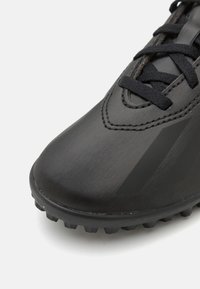 Primer plano de la parte frontal de una zapatilla deportiva negra texturizada con cordones negros y una suela de goma resistente, sobre un fondo blanco.