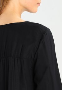 Blouse en tissu noir avec une coupe décontractée, présentant un col rond et des fronces subtiles au dos, offrant une texture lisse.
