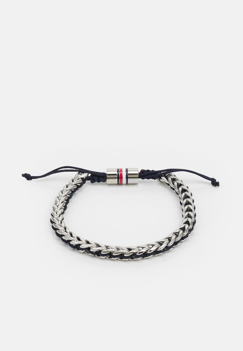 Bracelet en chaîne argentée avec un cordon bleu foncé, doté d'un fermoir cylindrique en métal avec des rayures rouges et blanches. Fermeture réglable à glissière.