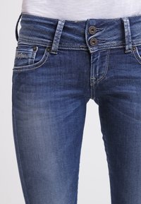 Jeans de mezclilla azul oscuro con cierre de doble botón, diseño de cinco bolsillos y un sutil desvanecimiento en los muslos. Presenta acentos de costura.