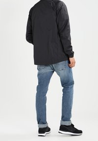 Svart lättviktsjacka, blå jeans och svarta skor; synlig sömnad på jeansen, uppvikta ärmar och vita skorselement.