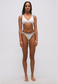 Γυναίκα με μακριά πλεξούδα στέκεται ξυπόλητη φορώντας λευκό σουτιέν και αντίστοιχα σλιπ με λωρίδα μέσης με λογότυπο, μπροστά σε απλό φόντο.