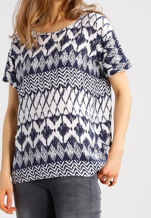 Femme portant un haut blanc à manches courtes avec des motifs géométriques et en zigzag bleu marine, assorti à un jean gris foncé ajusté.