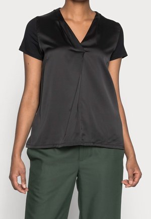 Nainen, joka pukee mustan, lyhythihaisen satiinipaidan, jossa on V-kaula-aukko, ja tummanvihreät, korkeavyötäröiset housut, seisoo yksinkertaisen taustan edessä.