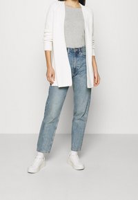 Bílý otevřený pletený cardigan přes šedé tričko, doplněný světle modrými džíny s vysokým pasem a bílými teniskami. Jednoduchý, ležérní outfit.