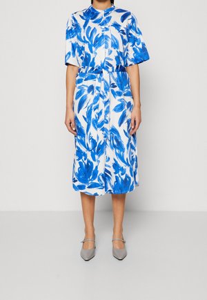 Femme portant une robe midi blanche avec des motifs floraux bleus en coups de pinceau, manches courtes, col haut et chaussures grises à bouts pointus avec des sangles.
