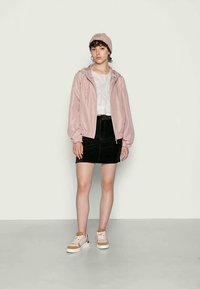Casaco bomber capuz rosa claro sobre uma blusa branca, combinado com uma saia mini preta. A modelo usa um gorro de malha rosa e ténis bege.