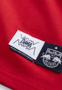 Rødt stof har to mærker: et hvidt rektangulært mærke med "PUMA" i sort og et marineblåt mærke med Red Bull Salzburg-logoet og tekst.