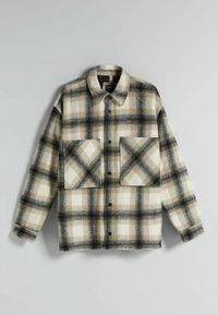 Camisa de lana de cuadros en tonos beige, blanco y negro, con botones en la parte delantera, dos grandes bolsillos frontales y un cuello clásico sobre un fondo liso.