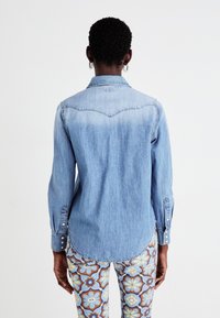 Camisa de ganga azul-claro com colarinho clássico, punhos abotoados e cintura curvada. Usada sobre calças florais estampadas com detalhes azuis e amarelos.