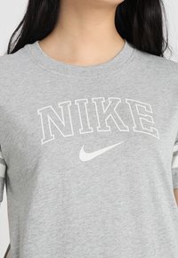 Camiseta gris de cuello redondo con el texto y logo "NIKE" en blanco. La tela parece suave con una textura jaspeada y acentos a rayas en las mangas.