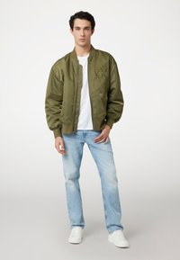 Jonge man die staat in een olijfgroene bomberjack, wit T-shirt, lichtblauwe jeans en witte sneakers tegen een effen achtergrond.
