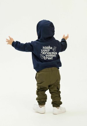Marinblå hoodie med ett grafiskt tryck på ryggen, kombinerad med olivgröna cargo-byxor och vita sneakers; mjukt tyg, avslappnad passform.