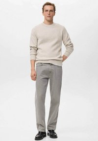Hombre con suéter beige de canalé, jeans gris claro y zapatos negros, de pie con una mano en el bolsillo contra un fondo blanco liso.