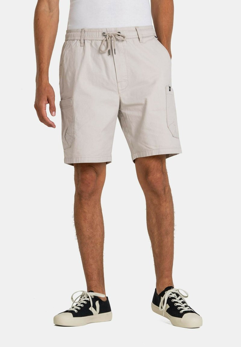 Pantalones cortos de carga de color beige claro con cintura elástica, cordón de ajuste y bolsillos laterales, combinados con zapatillas negras y blancas. Tejido texturizado.