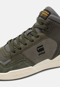 Valkoiset korkeavartiset tennarit oliivi vihreässä ja harmaassa nahassa. Ominaisuuksiin kuuluu teksturoitu pinta, nauhakuvaus ja logotieto sivussa.